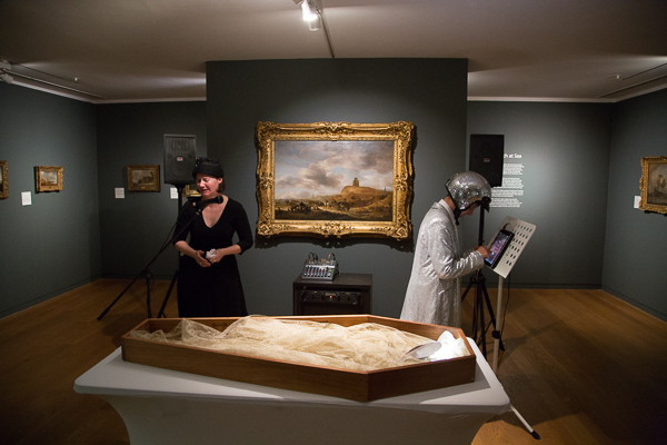 Performance of Space Adex and Manchester Art Gallery. A glittery DJ play music in front of an empty coffin. A mourner dressed in black cries into a microphone